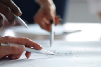 Close up of three people holding pens to a piece of paper on a desk.