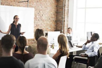 Woman standing at the front of a room presenting to a group of adults in an office environment.